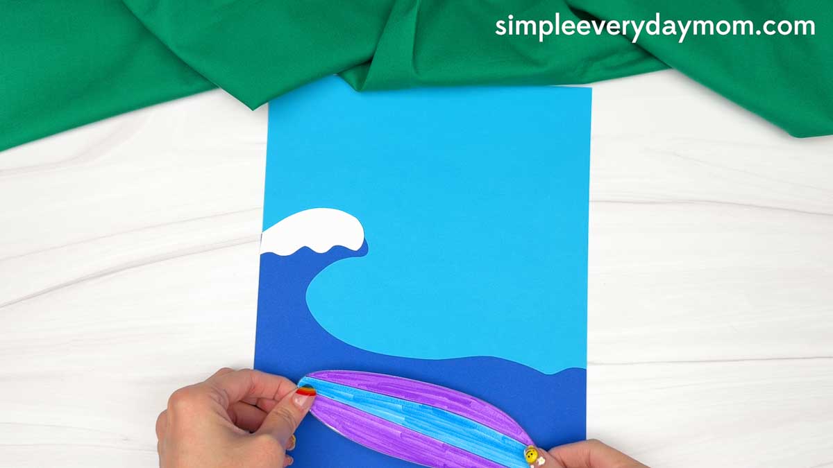 Hands positioning a colorful surfboard cutout on the ocean background for an elf surfing craft.