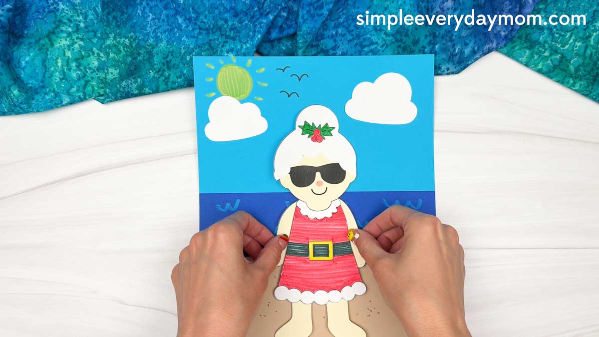 Hands positioning the Summer Mrs. Claus craft onto the completed beach scene background.