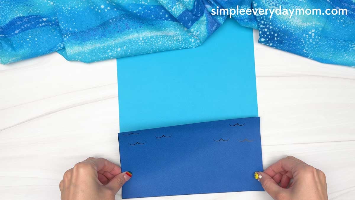 Hands hold blue cardstock paper layered to create an ocean scene for a Christmas in July gingerbread man craft.
