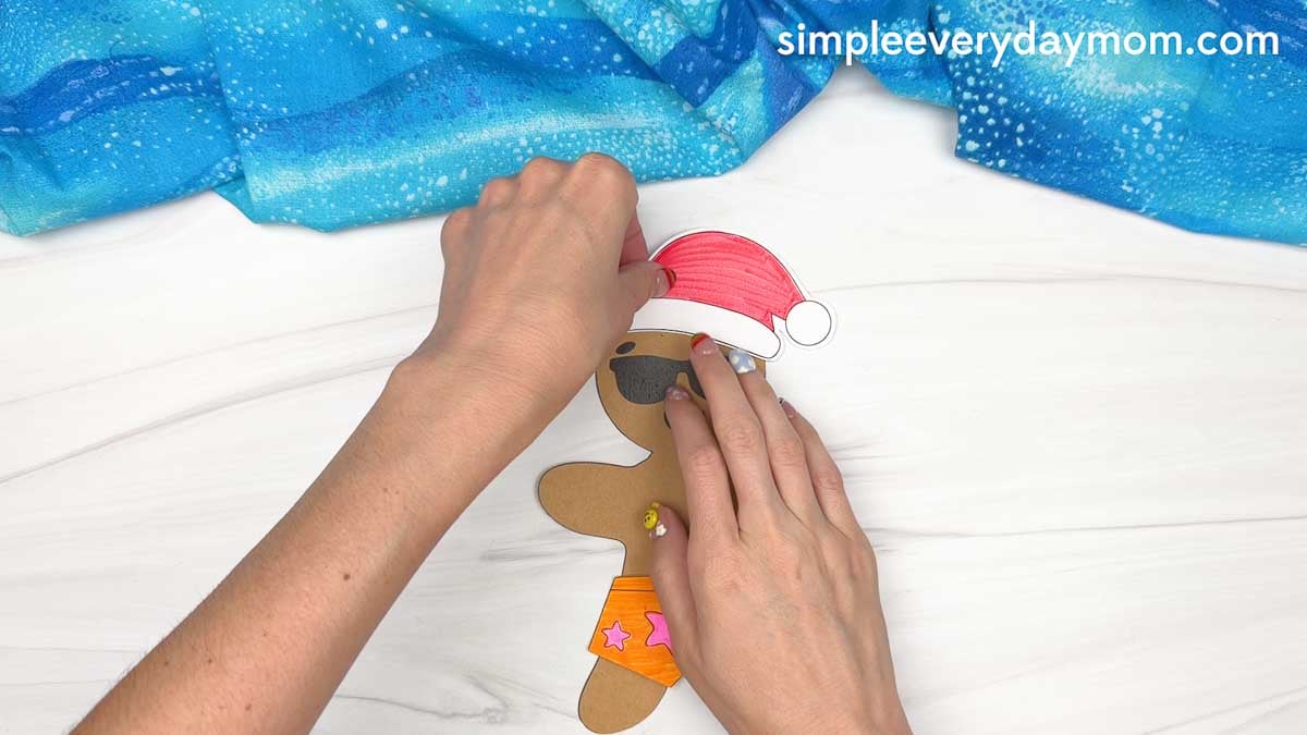 Hands place a red Santa hat on a beach-themed gingerbread man wearing sunglasses and orange swim shorts for a Christmas in July craft.