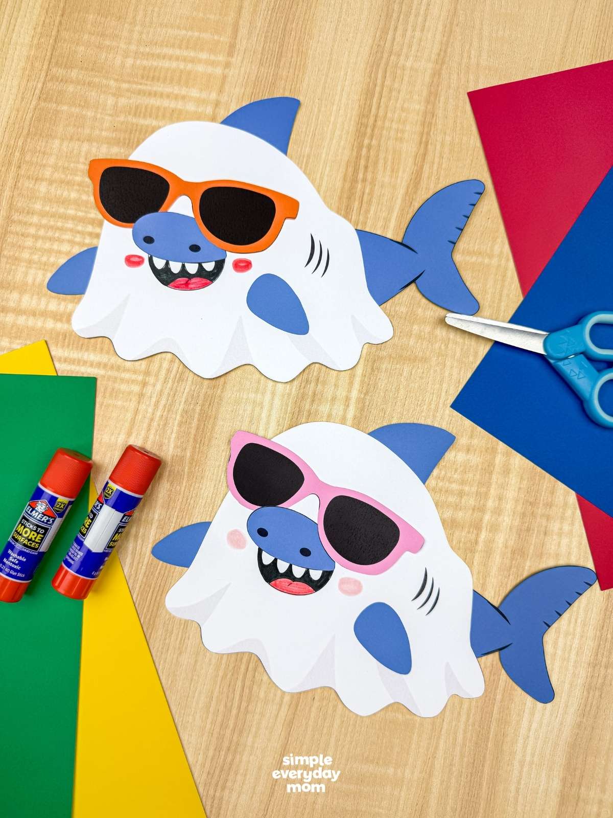 A completed craft of two Summerween ghost shark crafts with colorful sunglasses surrounded by paper, glue sticks, and scissors.