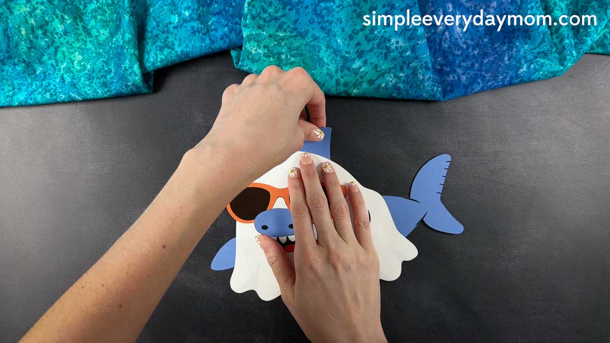 Hands attaching the blue dorsal fin to the top of a nearly completed Summerween ghost shark craft.
