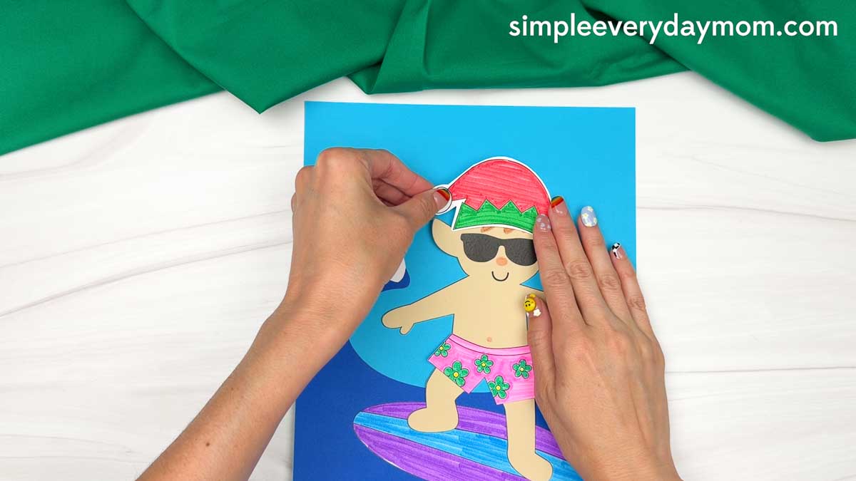 Hands attaching a red and green Santa hat to the surfing elf on a colorful surfboard ocean scene for an elf surfing craft.