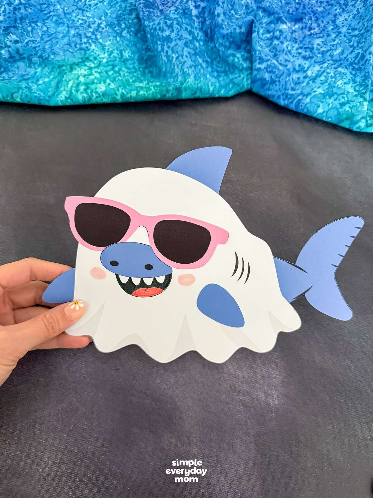 Hands holding a completed Summerween ghost shark craft with pink sunglasses on a dark background.