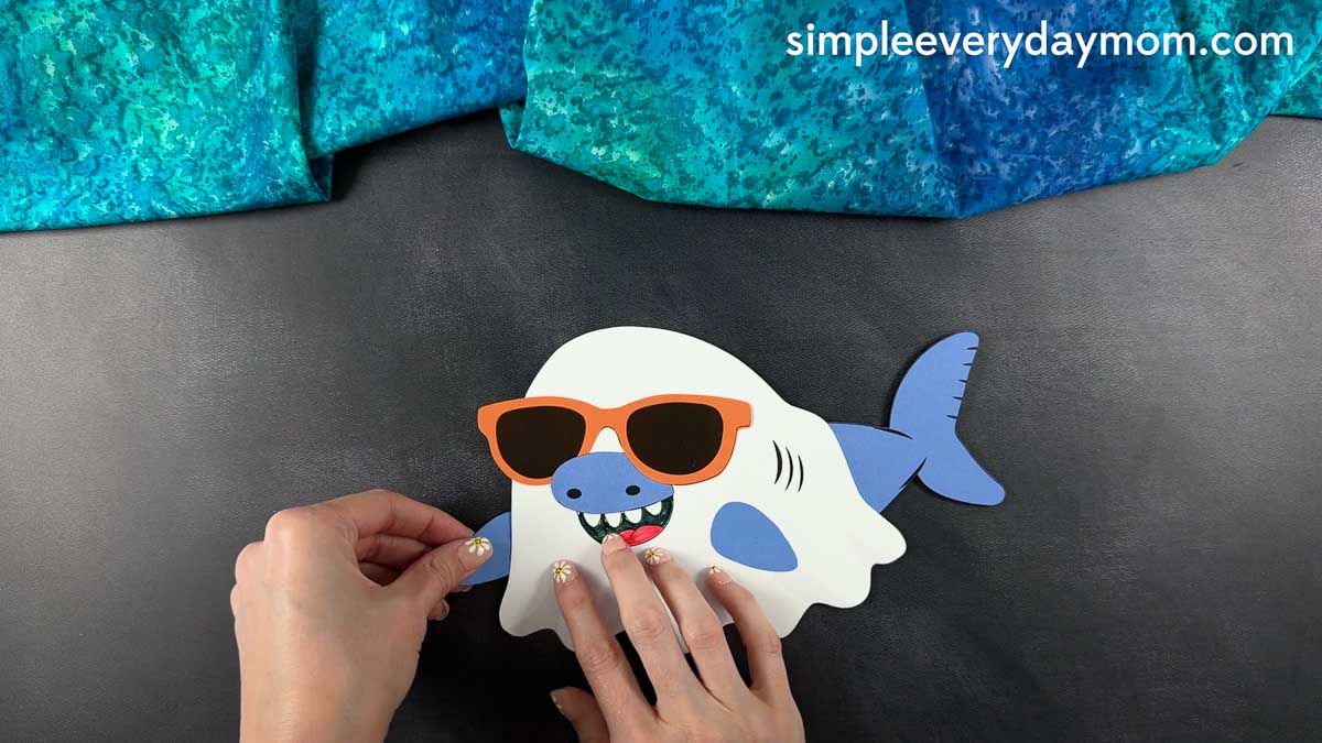 Hands attaching the final blue shark fin to a nearly completed Summerween ghost shark craft with sunglasses.