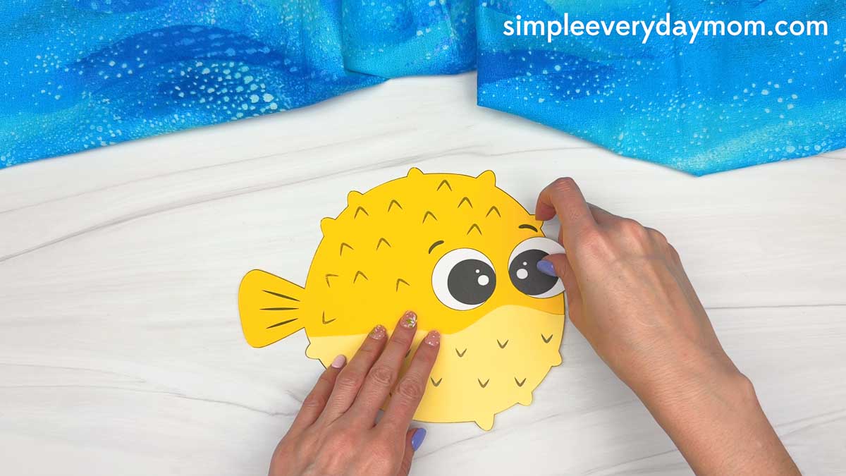 Hands attaching a large eye piece to a yellow pufferfish paper craft during assembly.