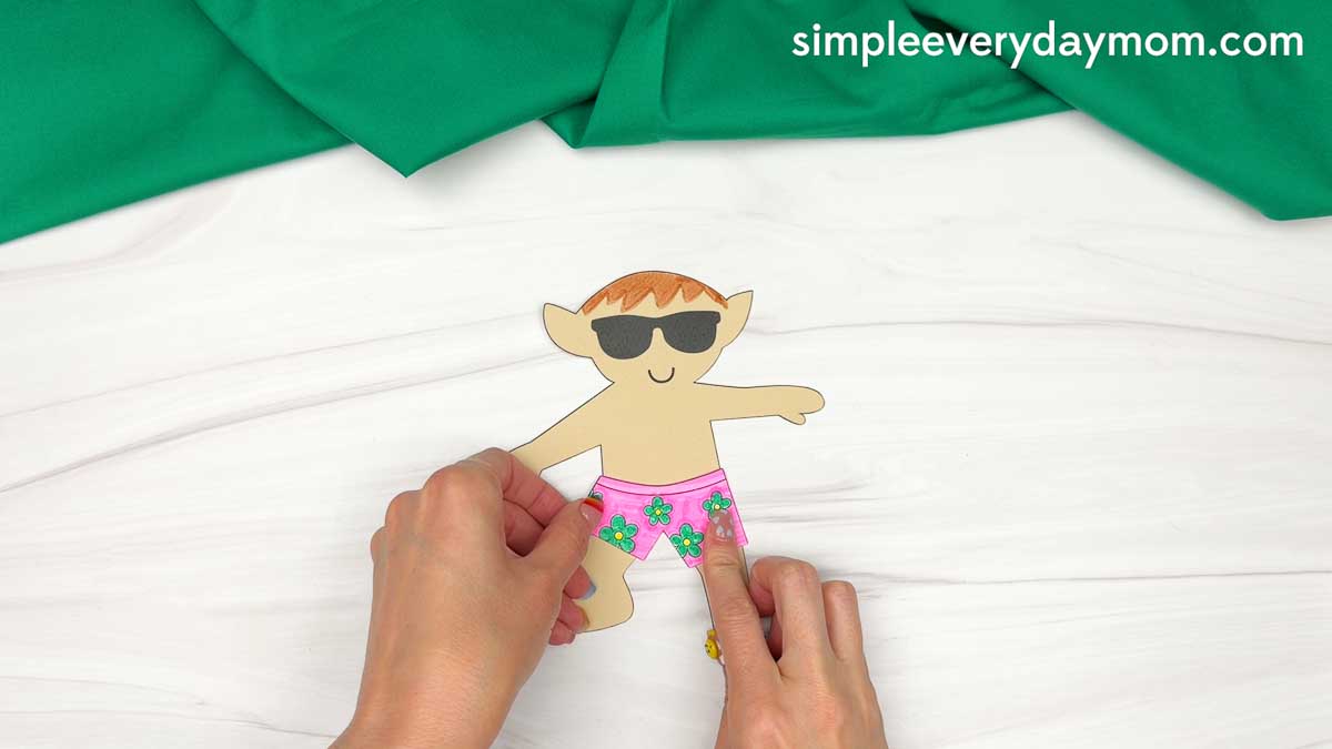 Hands attaching pink floral swim trunks to a cut out elf wearing sunglasses for an elf surfing craft.