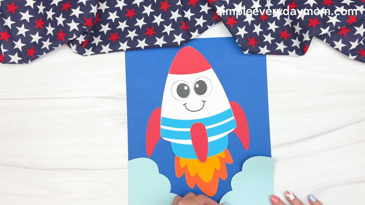 A completed 4th of July rocket craft on blue cardstock with paper clouds and colorful flame exhaust creating a rocket launch scene.