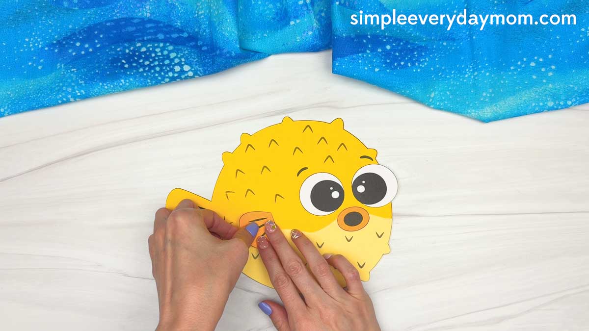 Hands attaching the side fin to a yellow pufferfish paper craft with large eyes and a small round mouth.