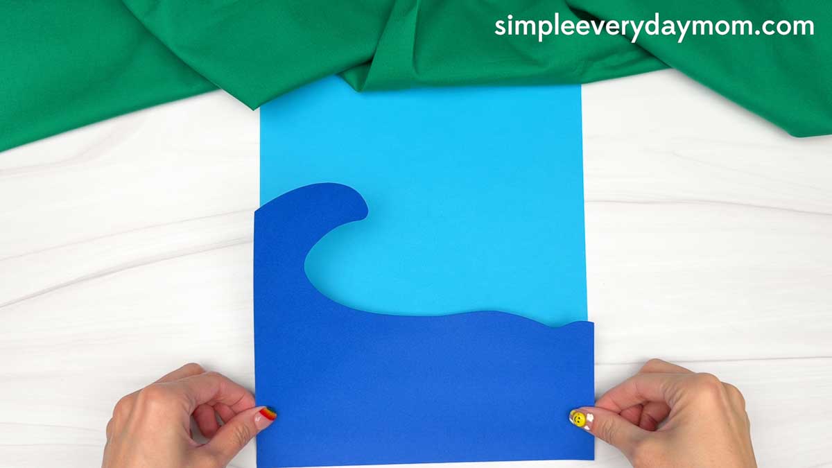 Hands holding blue cardstock waves layered over light blue paper for the ocean background of an elf surfing craft.