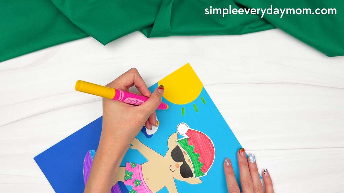 Hands drawing sun rays around a yellow paper sun above the surfing elf craft scene.