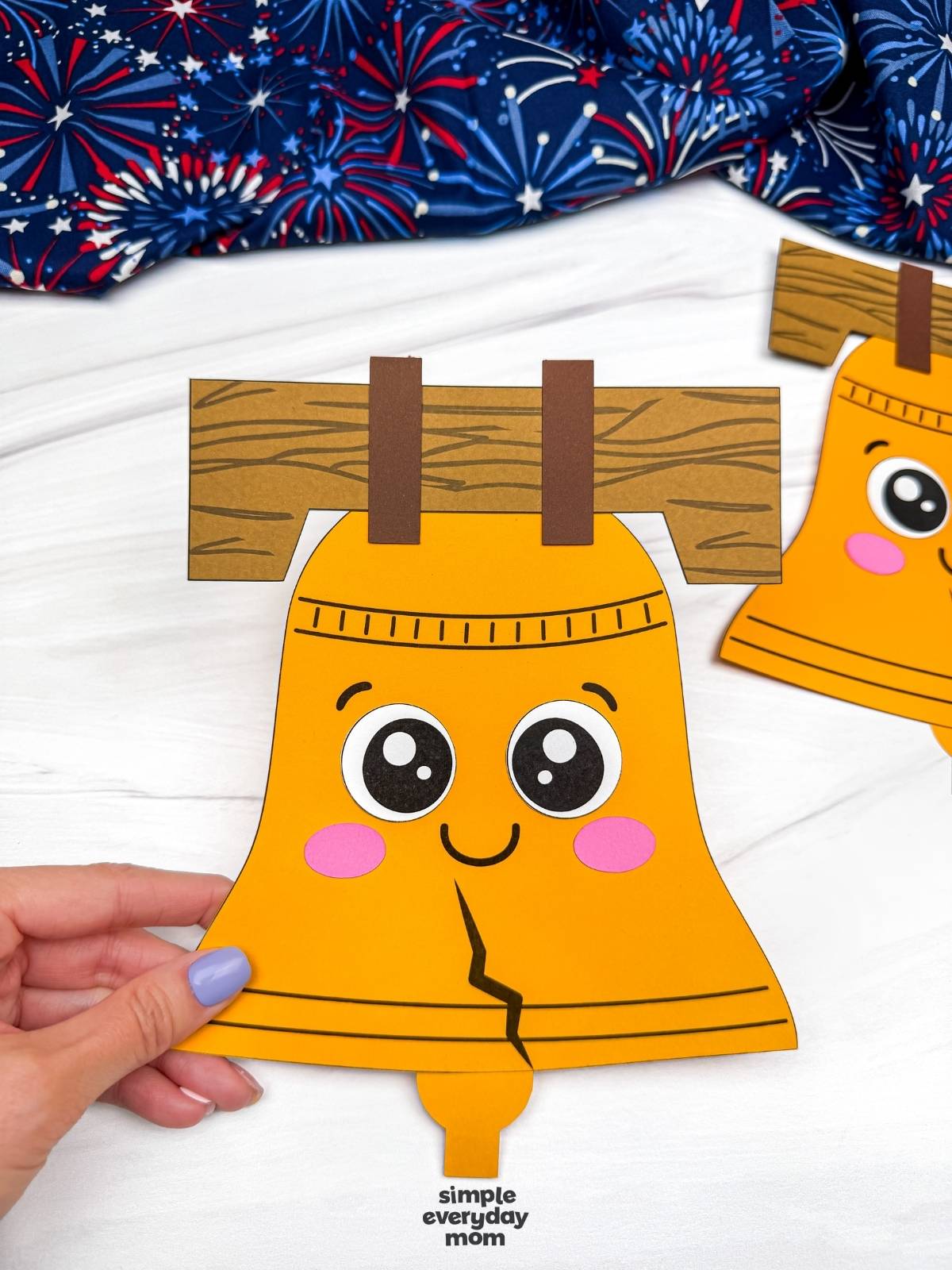Hands holding a completed Liberty Bell paper craft with a smiling face, cracked bell design, and wood beam support.