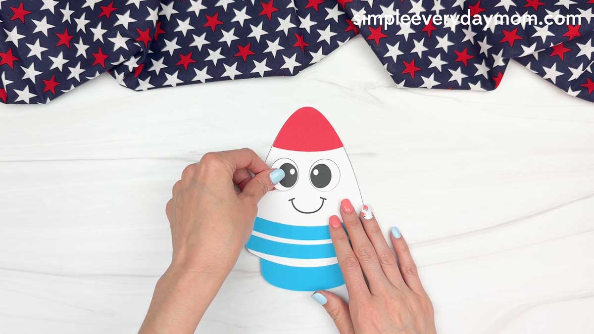Hands attaching large paper eyes to a 4th of July rocket craft on a white tabletop.