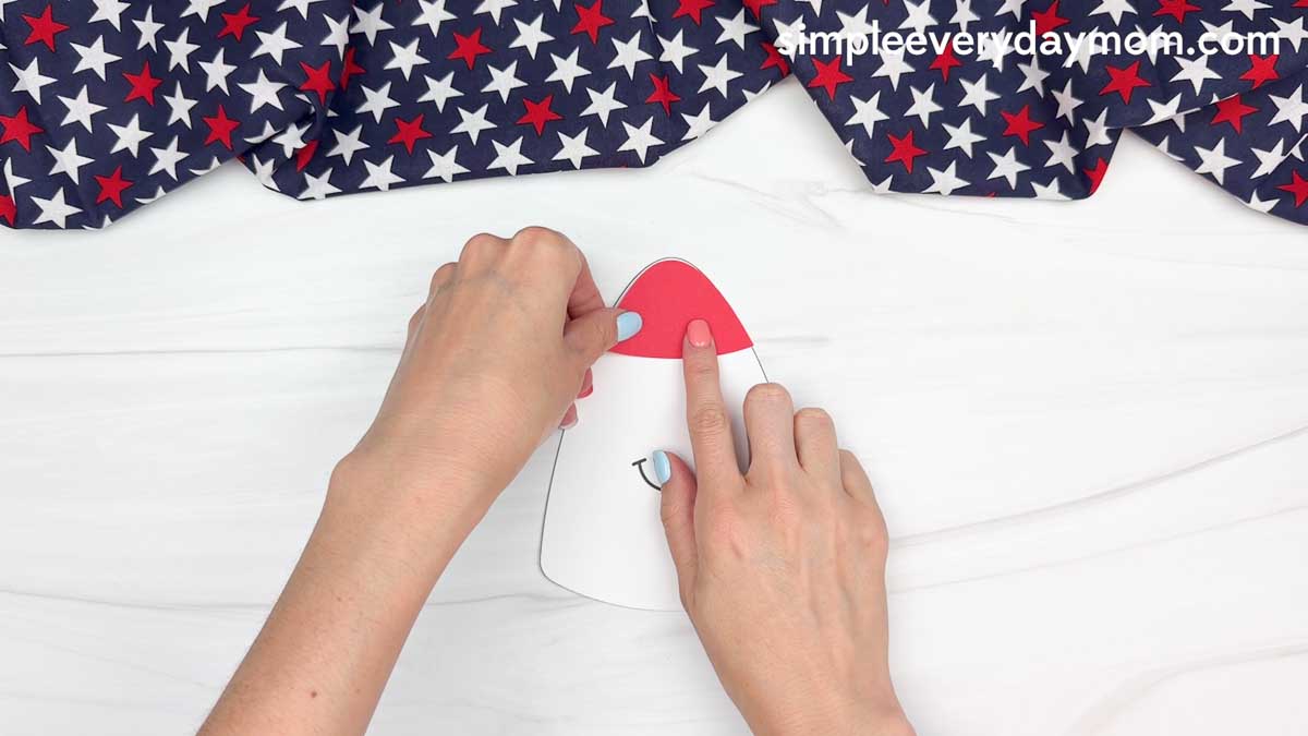Hands attaching a red paper rocket top to a 4th of July rocket craft on a white tabletop.