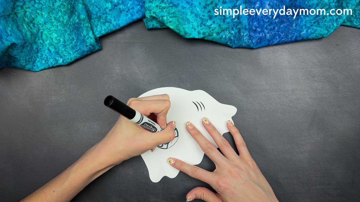 Hands drawing the ghost shark's mouth details on a white craft cutout with a black marker.