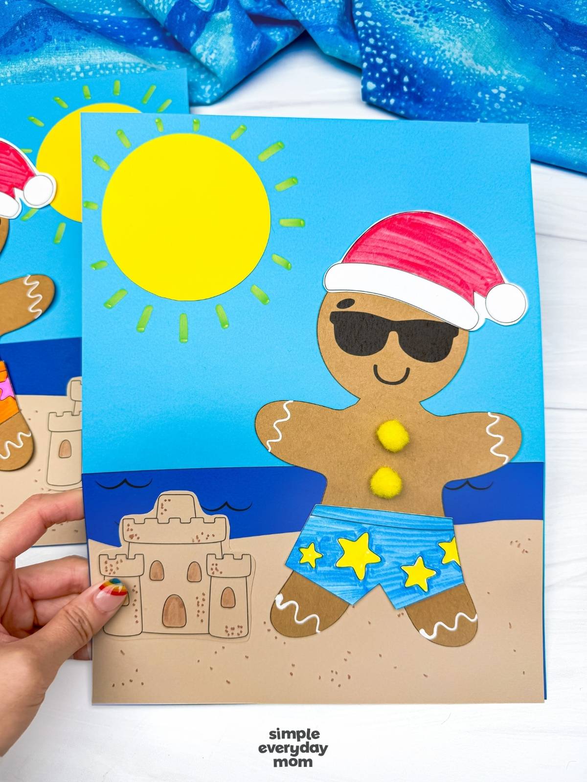 Hands hold a completed Christmas in July gingerbread beach craft featuring a gingerbread man in blue star swim shorts, sunglasses, a Santa hat, and a sandy beach scene with a sandcastle.