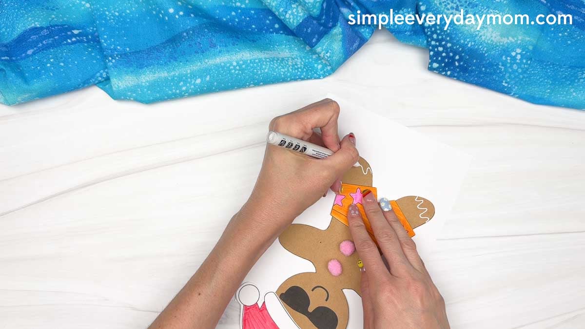 Hands draw white icing details on a beach-themed gingerbread man with pink buttons, orange swim shorts, sunglasses, and a Santa hat for a Christmas in July craft.