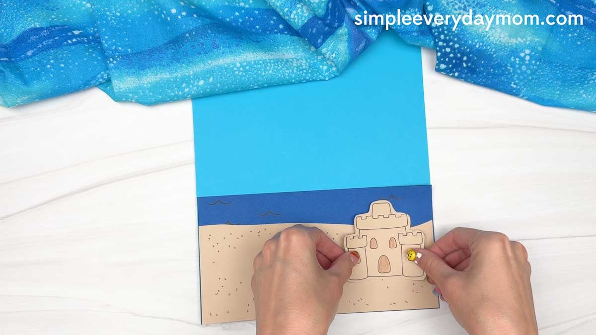Hands place a paper sandcastle cutout onto a beach scene background for a Christmas in July gingerbread man craft.