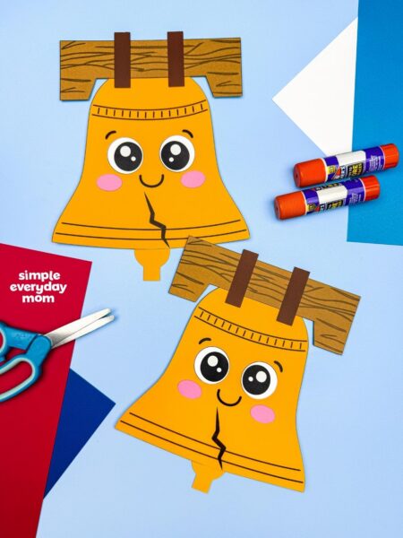 A completed Craft featuring two smiling Liberty Bell paper crafts displayed with scissors, glue sticks, and colorful paper.