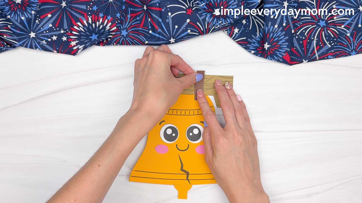 Hands attaching a paper strap to the wood beam on a Liberty Bell paper craft.