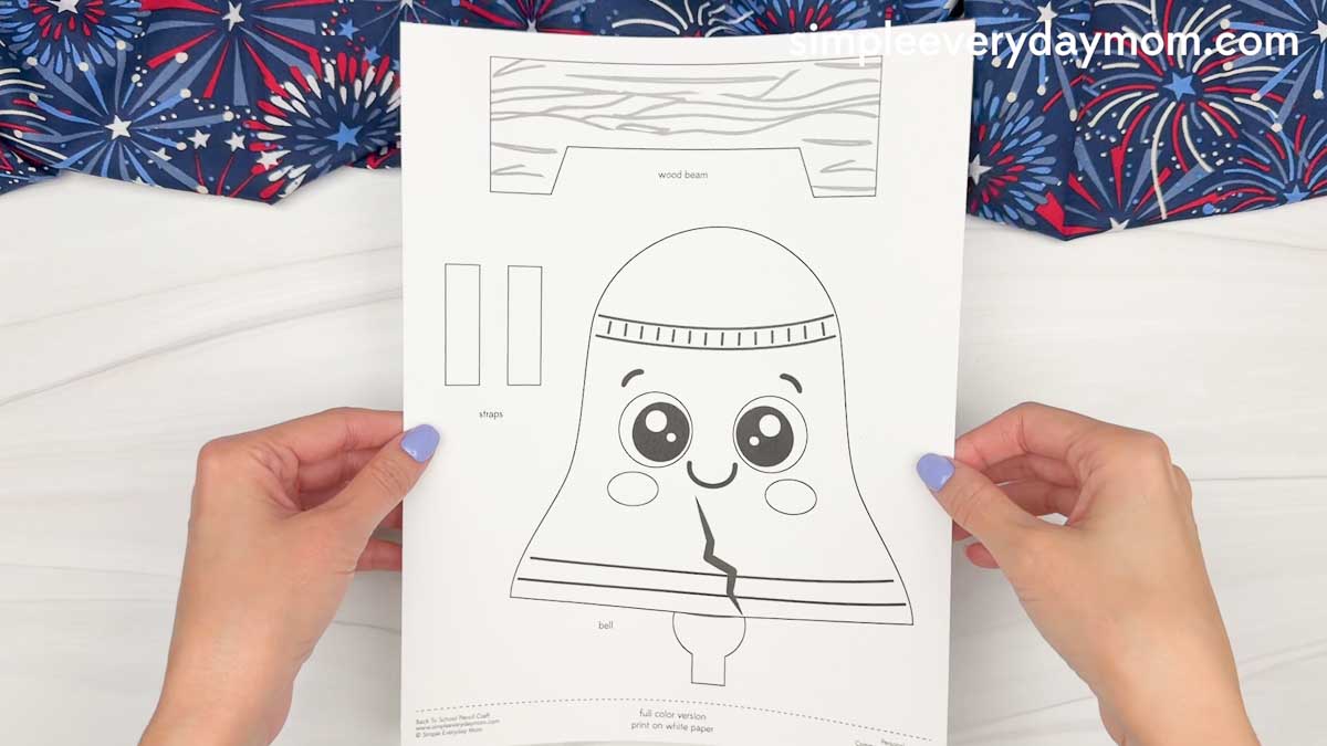 Hands holding a printed Liberty Bell paper craft template with separate wood beam and strap pieces ready to cut out and assemble.
