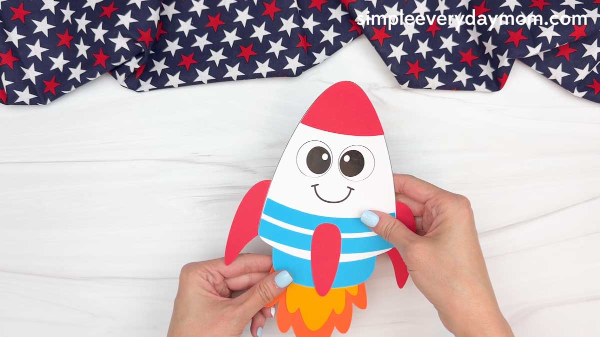 Hands holding a completed 4th of July rocket craft with colorful flame exhaust on a white tabletop.