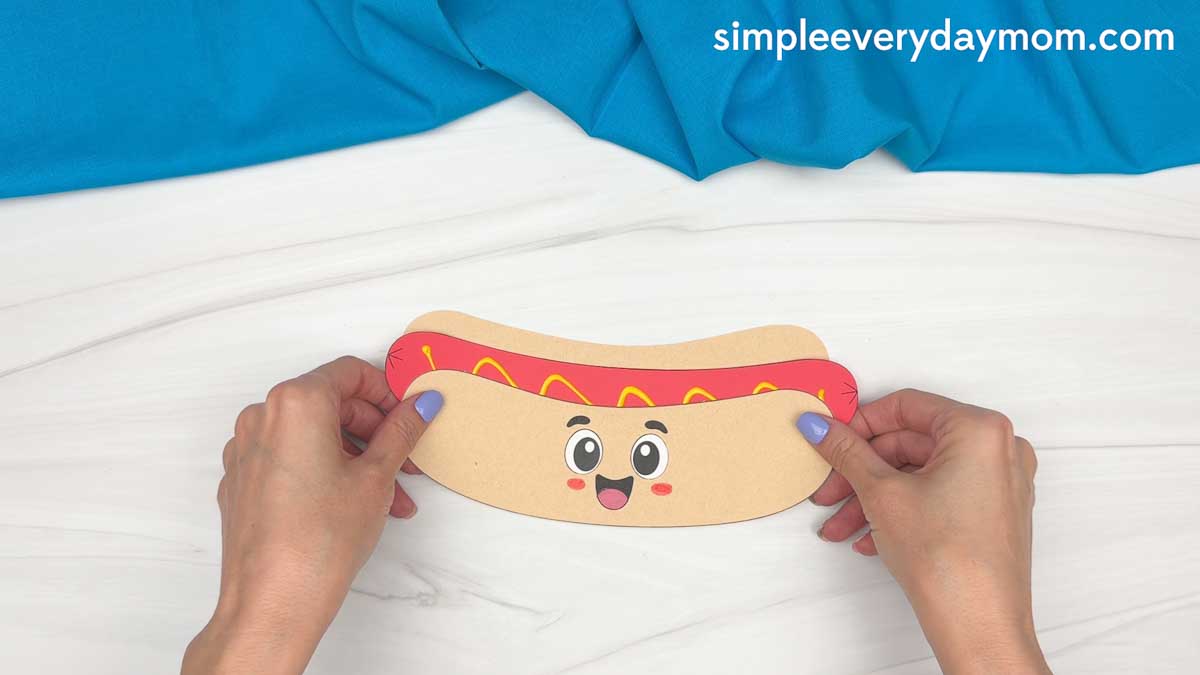 Hands attaching the hot dog cutout to the back bun piece to assemble the hot dog craft.