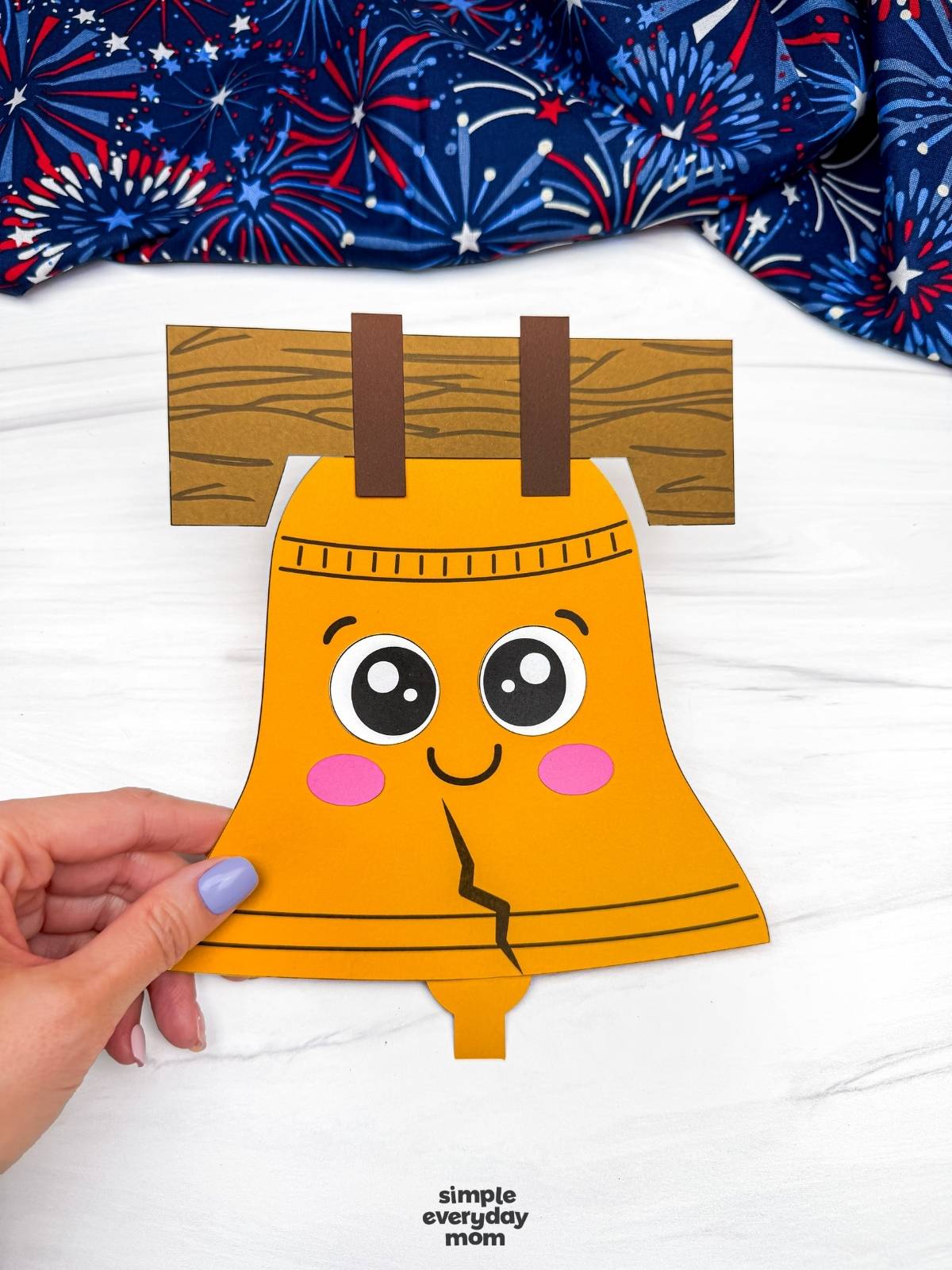Hands holding a completed Liberty Bell paper craft with a smiling face and wood beam hanger.