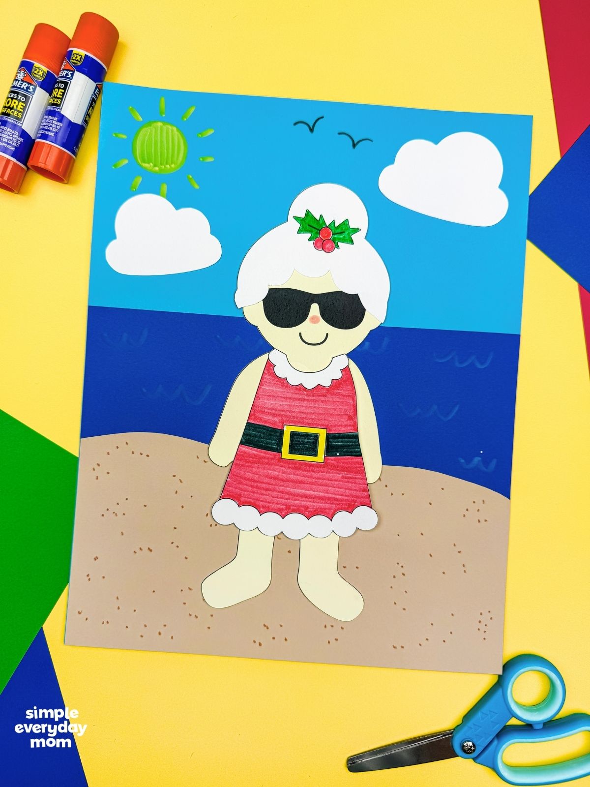 A completed craft of Summer Mrs. Claus on a beach scene with colorful paper, glue sticks, and scissors.
