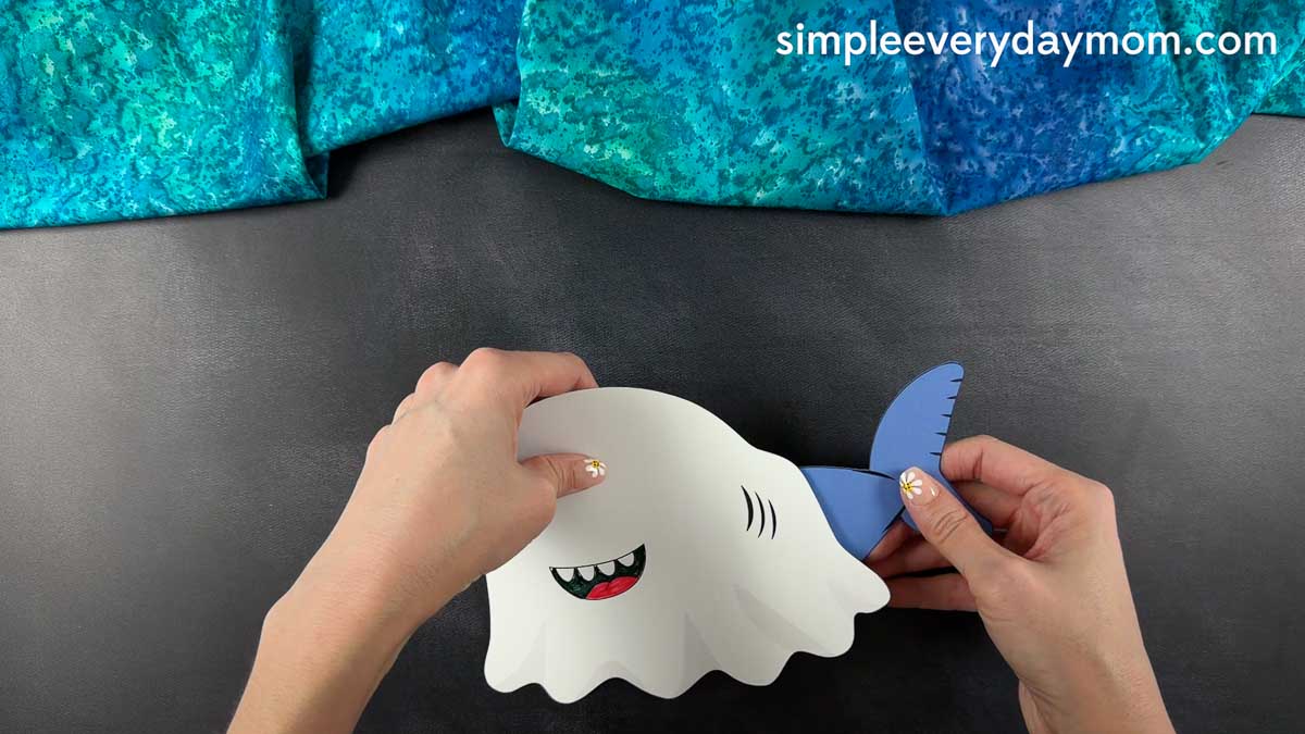Hands attaching a blue shark tail to a white ghost shark craft during assembly.
