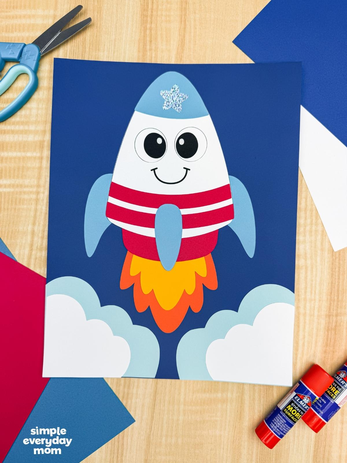 A completed 4th of July rocket craft launch scene with patriotic colors, fluffy paper clouds, colorful flame exhaust, and craft supplies.