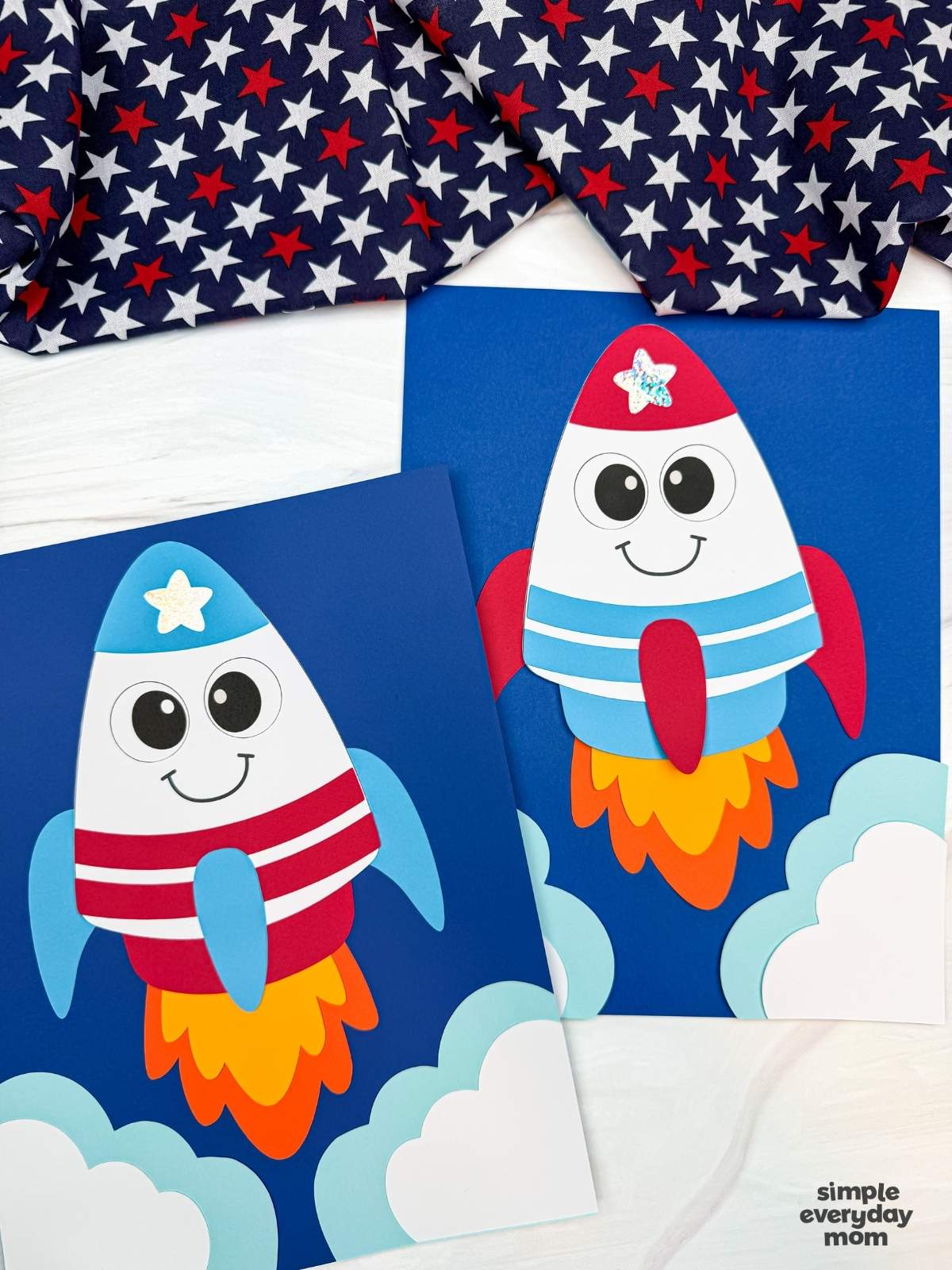 A completed 4th of July rocket craft display featuring two patriotic paper rocket launch scenes with clouds, colorful flame exhaust, and blue cardstock backgrounds.