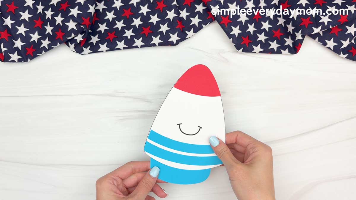 Hands holding a partially assembled 4th of July rocket craft with a red top and blue stripes on a white tabletop.