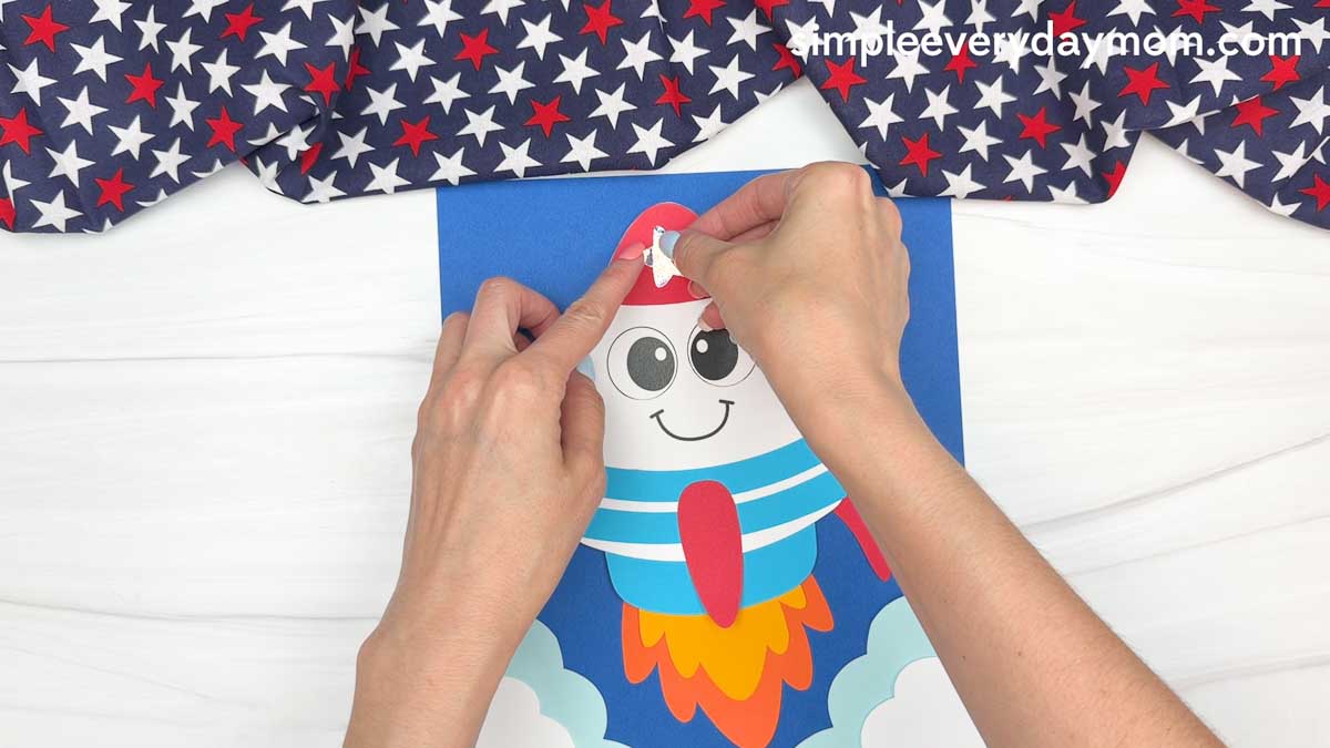 Hands adding a star decoration to a completed 4th of July rocket craft launch scene with clouds and colorful flame exhaust.
