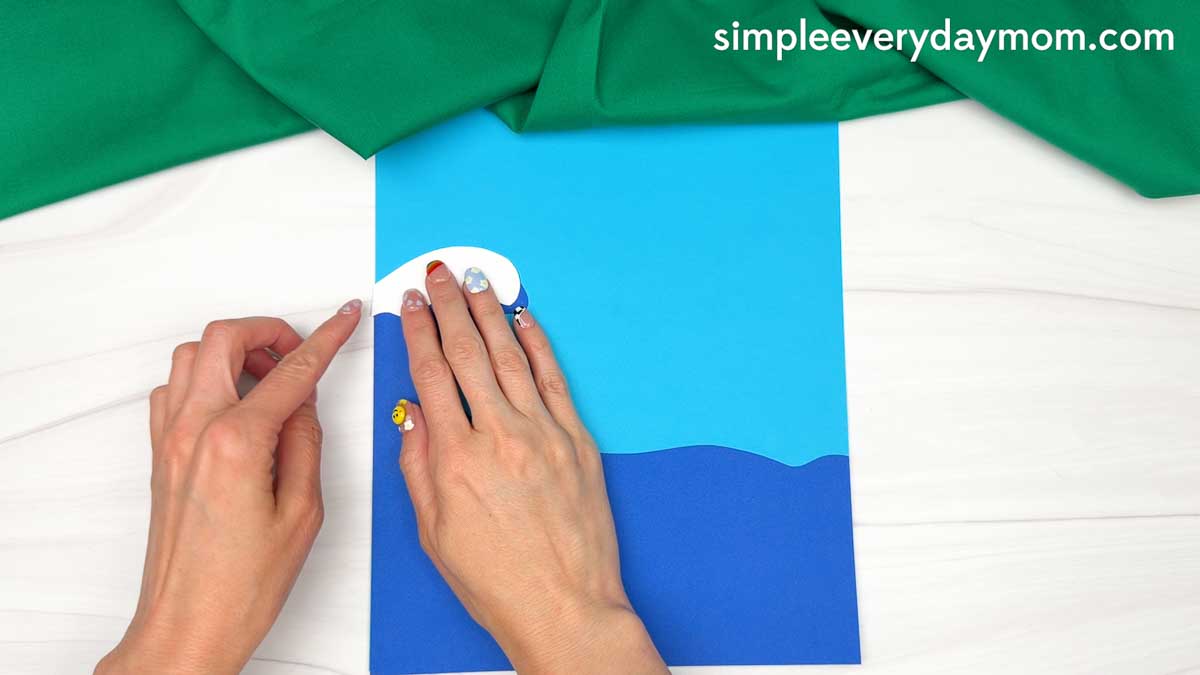 Hands placing a white foam wave crest on blue paper waves to create the ocean scene for an elf surfing craft.