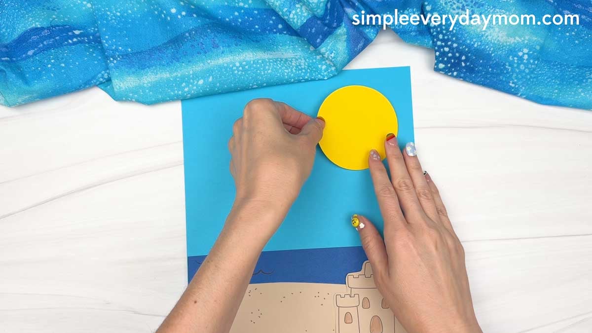 Hands place a yellow paper sun onto a beach scene background with ocean waves and a sandcastle for a Christmas in July gingerbread man craft.