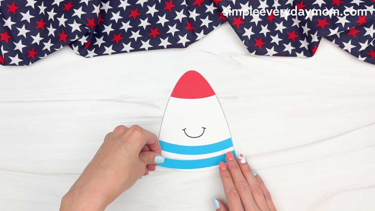 Hands adding blue paper stripes to a 4th of July rocket craft on a white tabletop.