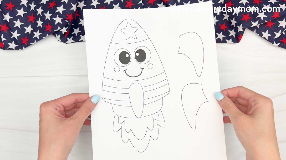 Hands holding a printable 4th of July rocket craft template with a smiling rocket and separate cut out fin pieces on a white background.