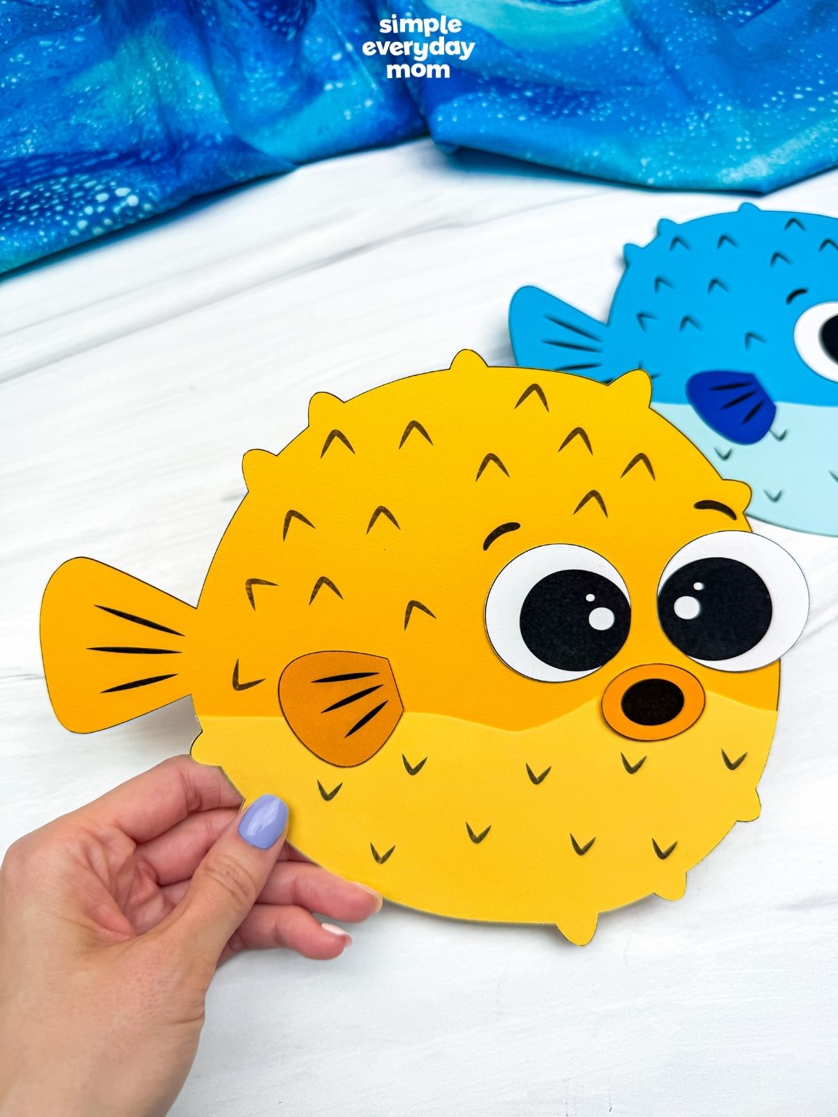 Hands holding a completed yellow pufferfish paper craft with a blue pufferfish craft in the background.
