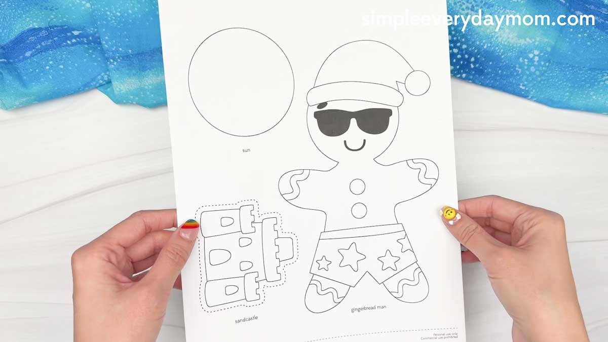 Hands hold a printable Christmas in July gingerbread man craft template with a sun, sandcastle cutout, and beach-themed gingerbread character wearing sunglasses and a Santa hat.