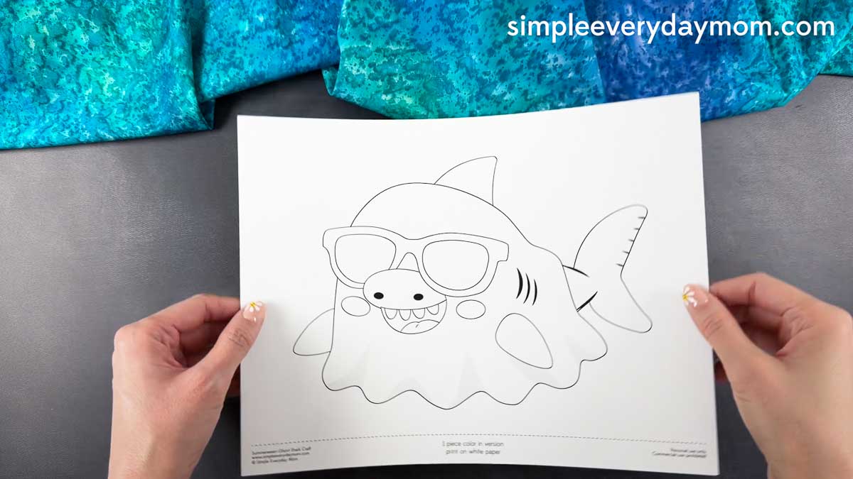 Hands holding a printable Summerween ghost shark craft template with sunglasses on a gray background.