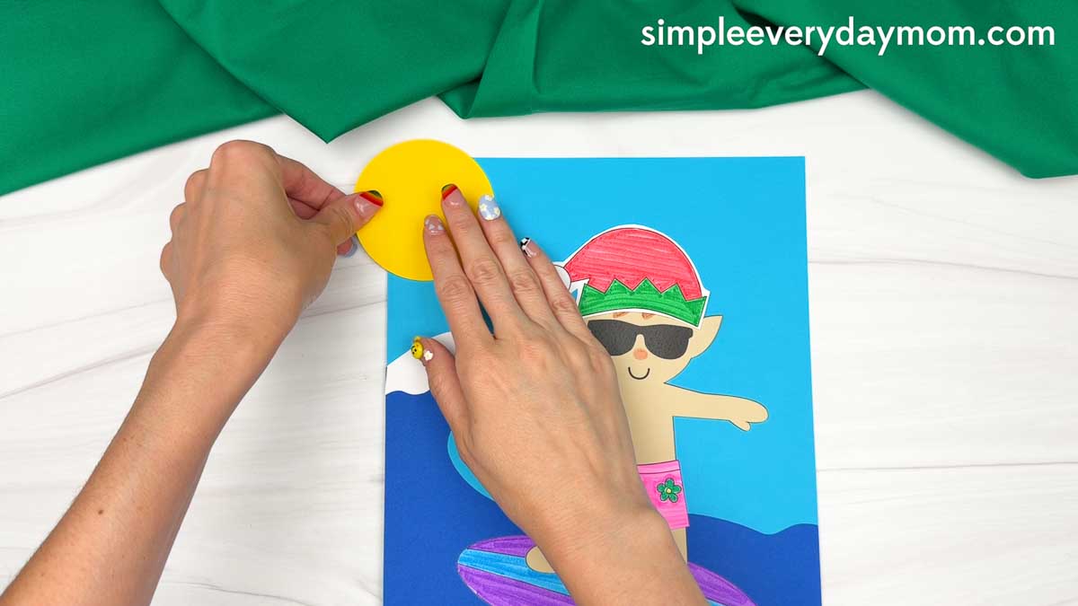 Hands placing a yellow paper sun onto the ocean background above the surfing elf craft scene.