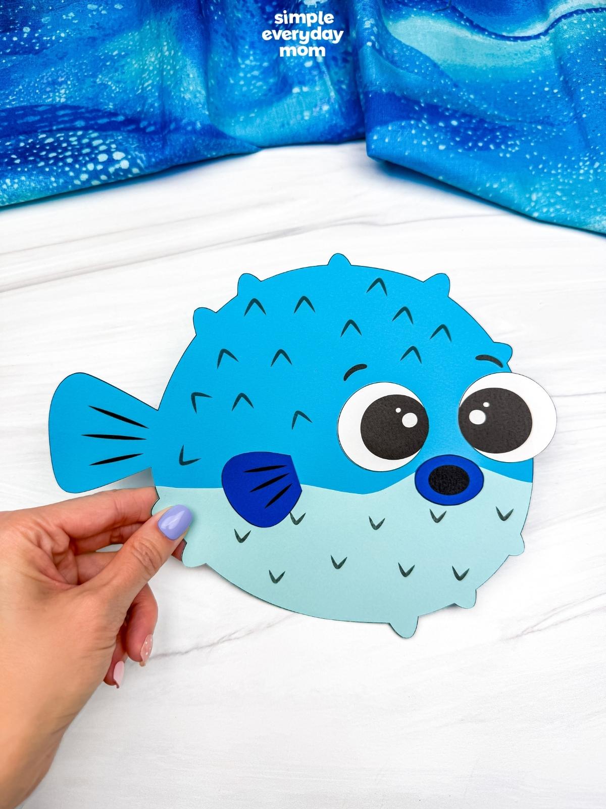 Hands holding a completed blue pufferfish paper craft with large eyes on a white tabletop.