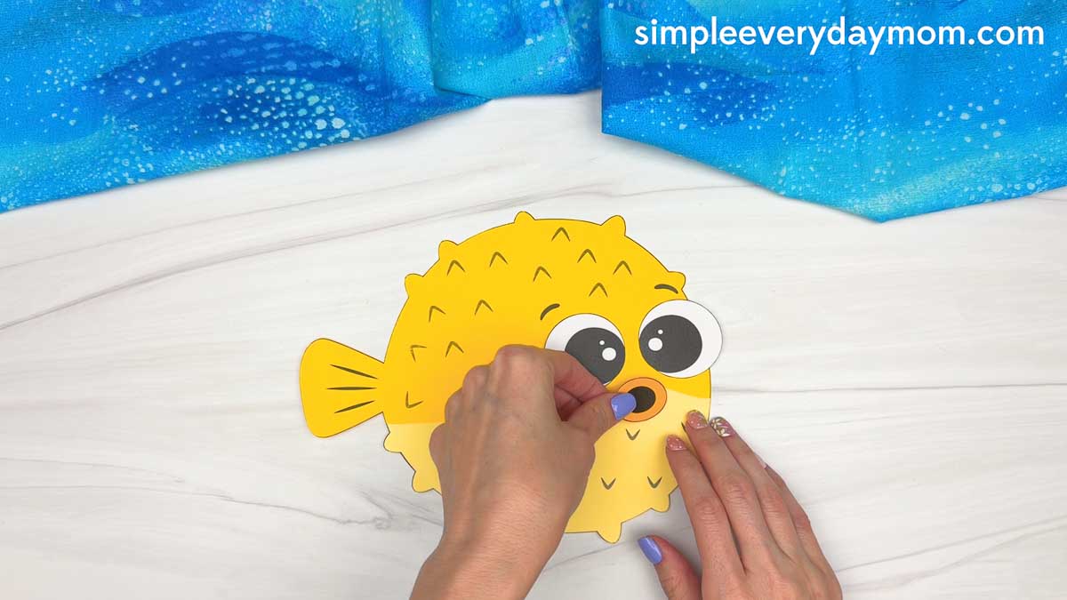 Hands attaching the pufferfish mouth to a yellow paper craft with large googly eyes.