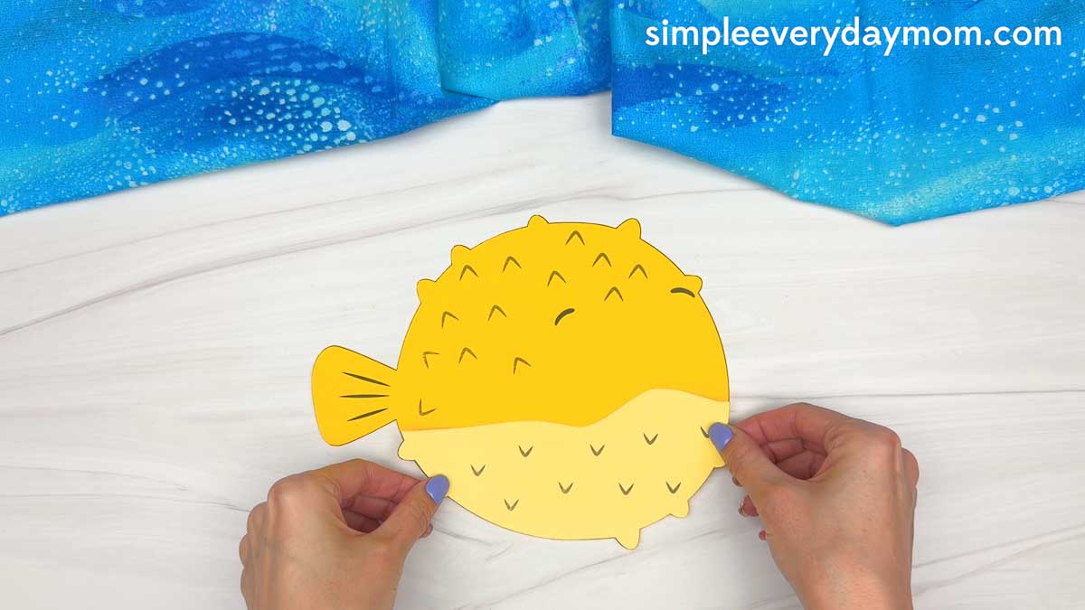 Hands holding a partially assembled yellow pufferfish paper craft with the body pieces attached.