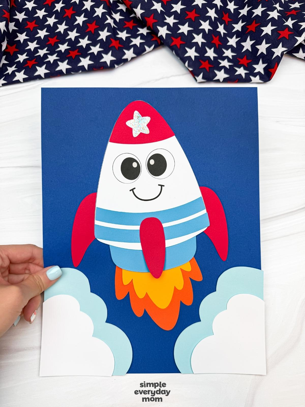 Hands holding a completed 4th of July rocket craft launch scene with paper clouds, colorful flame exhaust, and a blue cardstock background.