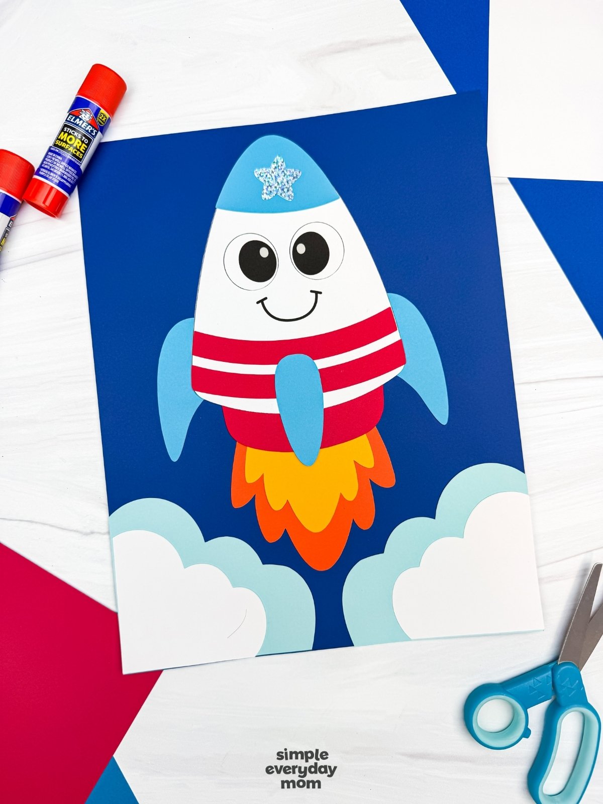 A completed 4th of July rocket craft launch scene with patriotic colors, paper clouds, colorful flame exhaust, and craft supplies arranged around the project.