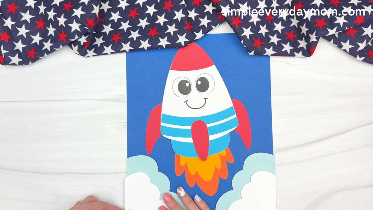 A completed 4th of July rocket craft launch scene with fluffy paper clouds, colorful flame exhaust, and a blue cardstock background.