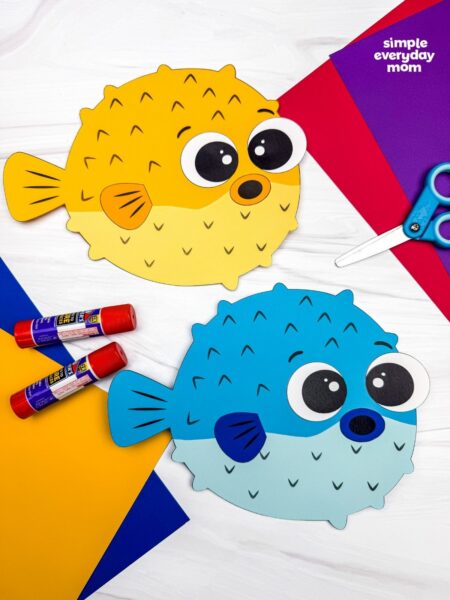 A completed craft featuring yellow and blue pufferfish paper crafts displayed with colorful paper, glue sticks, and scissors.