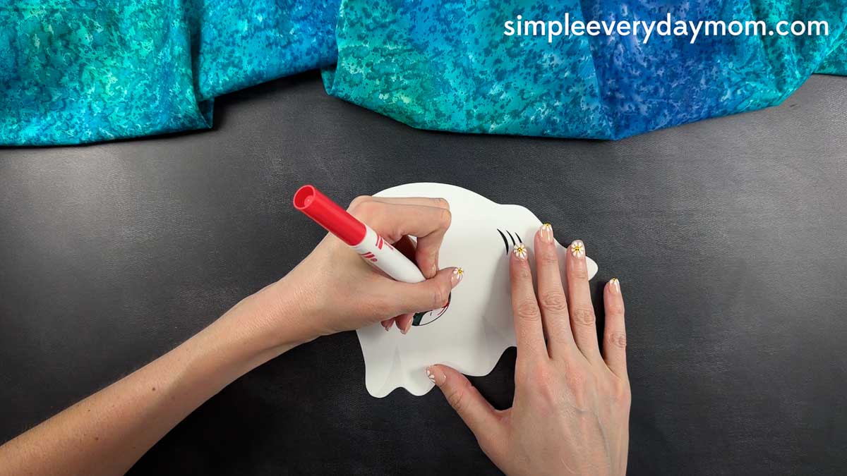 Hands tracing facial details onto a white ghost shark craft cutout with a red marker.
